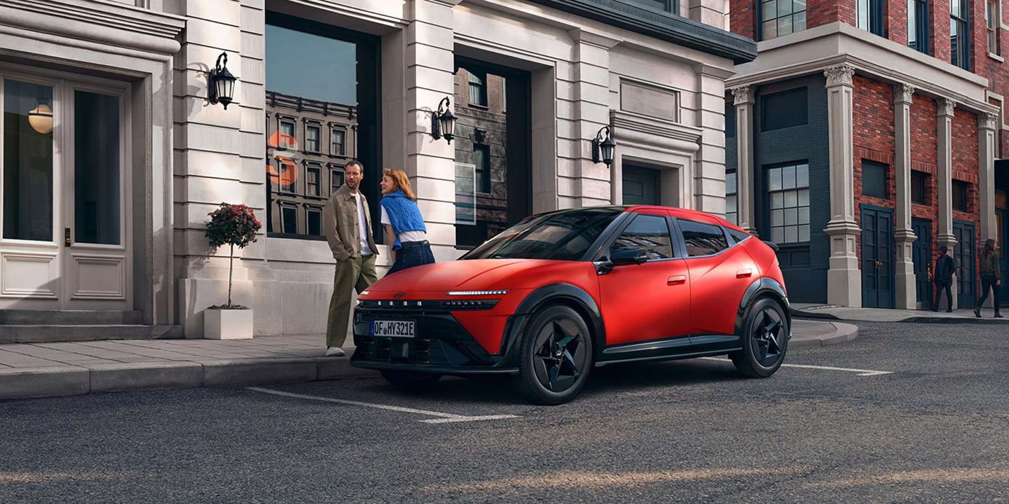 En rød crossover-SUV er parkeret på gaden foran en bygning, og en mand og en kvinde står sammen på det nærliggende fortov.