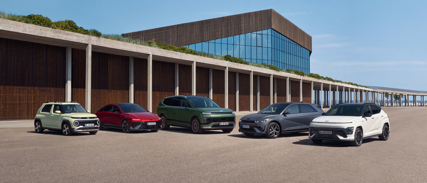 Fem forskellige Hyundai-elbiler står parkeret på rad og række uden for en moderne glas- og træbygning på en solrig dag.