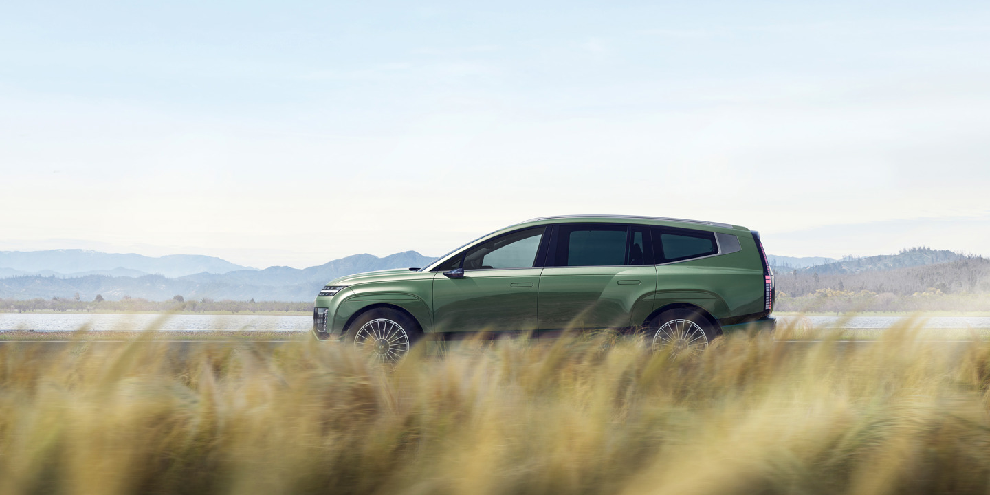 En grøn IONIQ 9 kører på en vej, omgivet af højt græs i forgrunden og bakker i baggrunden under en klar himmel.