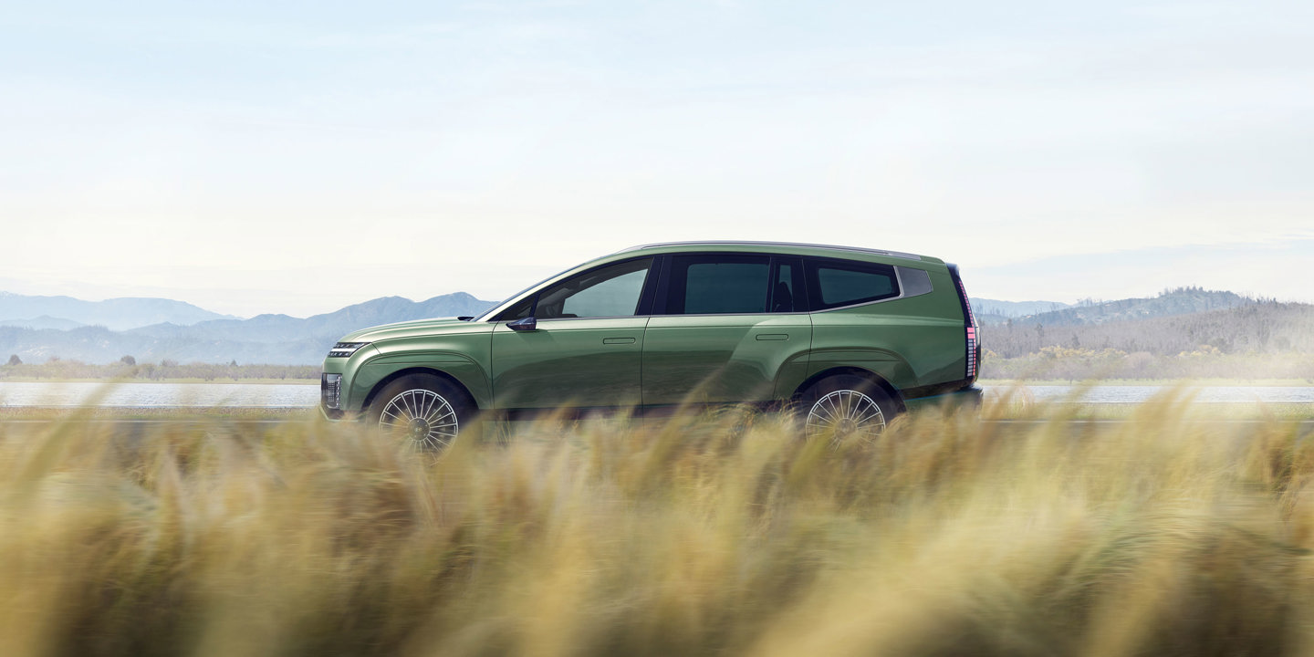 En grøn Hyundai IONIQ 9 kører på en vej ved siden af højt græs, med majestætiske bjerge og en smuk skyet himmel i baggrunden.