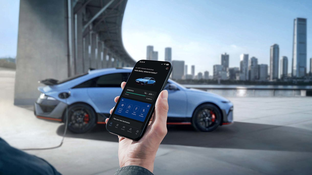 En person holder en smartphone foran en blå bil, der er parkeret udendørs i nærheden af en moderne arkitektonisk struktur.