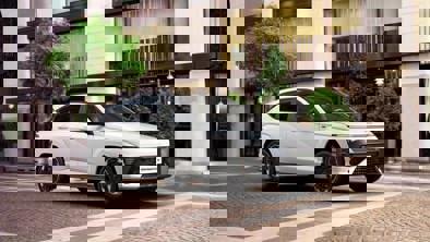 En hvid kompakt SUV parkeret på en brostensbelagt gade foran en bygning med træer og balkoner.