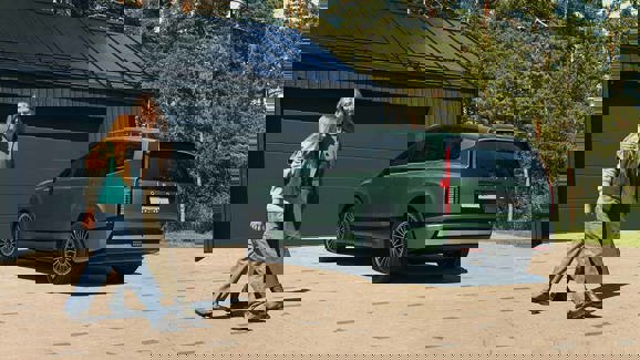 En person lufter en hund i en indkørsel foran en sort SUV og et hus med en garage omgivet af træer.