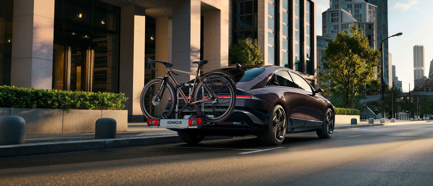 En elegant sort bil med to cykler på bagagebæreren er parkeret på en gade i byen, der er omgivet af moderne bygninger og træer.