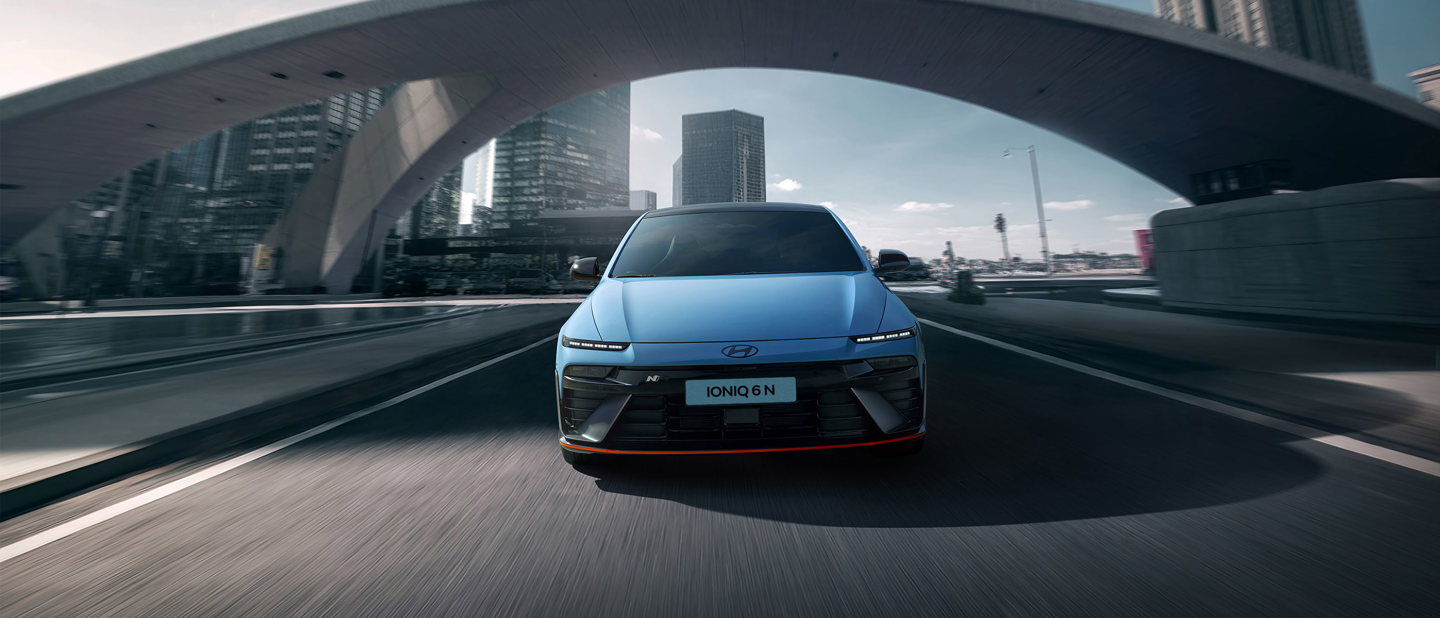 En blå Hyundai Ioniq 6 N kører på en gade i byen, omgivet af moderne skyskrabere og en stor buet bro.