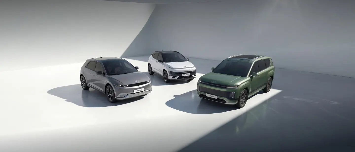 Tre moderne elbiler - grå, hvid og grøn - er parkeret i et slankt, stærkt oplyst indendørs rum med glatte, buede hvide vægge.