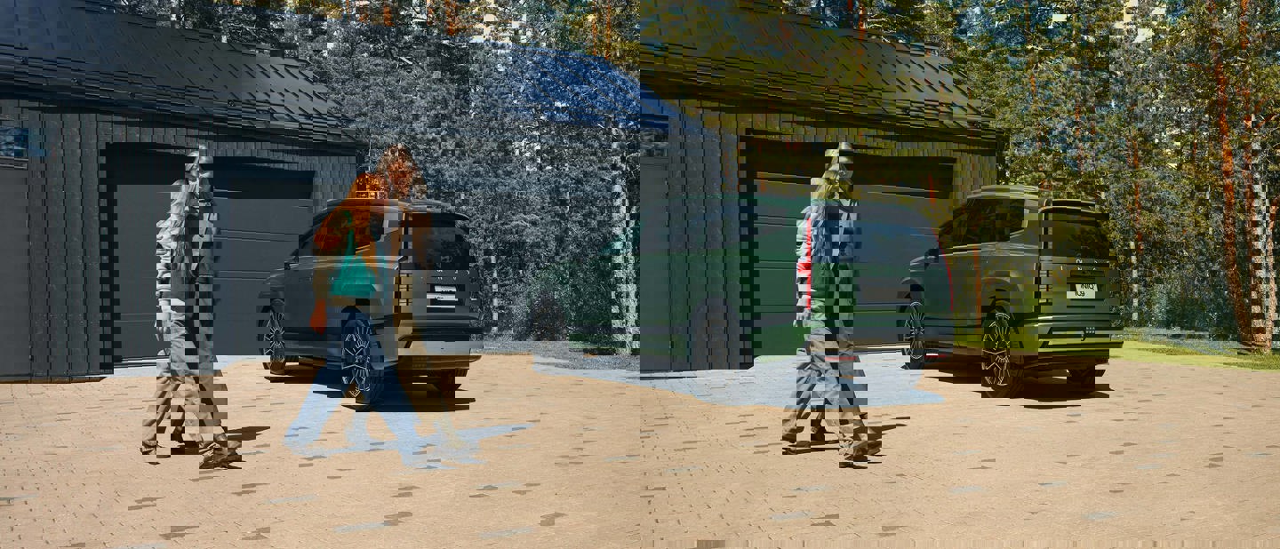 En person går forbi en grå SUV, der er parkeret i en indkørsel foran et moderne hus med træer i baggrunden.