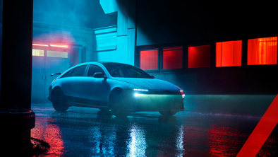 En slank, moderne bil med forlygterne tændt er parkeret i et vådt, neonoplyst bymiljø om natten.