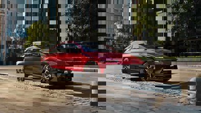 En rød bil parkeret på en brostensbelagt gade i en by med bygninger og træer i baggrunden. Billedet virker digitalt forvrænget.